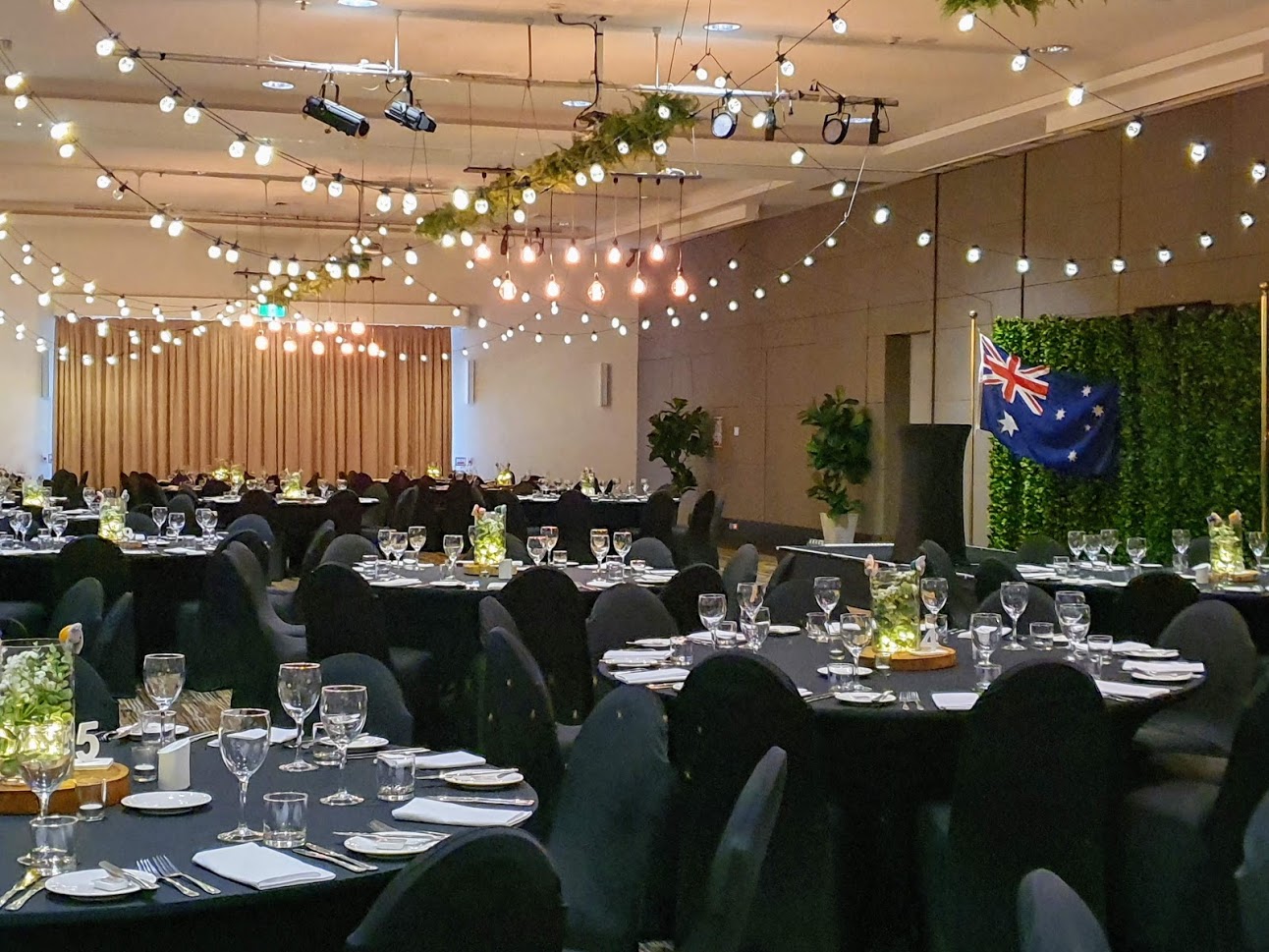Jetty Harbour Marina Rooms - Marquee ceiling canopy, greenery & Festoon lights, Edison chandeliers, Black chair covers, Table decoration glass vase  battery fairy lights, eucalyptus, babies breath on a timber log- Pacific Bay Resort