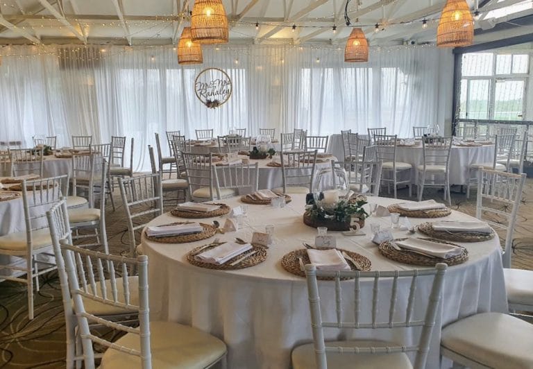 White fabric backdrop, waterfall fairy light curtain, festoon ceiling lighting, white round tables, white Tiffany chairs, Rattan placemats, Glass lotus centre piece with silk ivy and timber round plate, table numbers, tealights