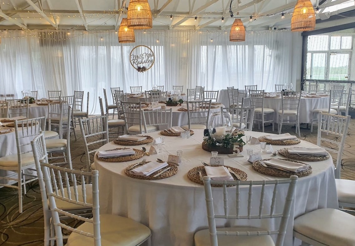 White fabric backdrop, waterfall fairy light curtain, festoon ceiling lighting, white round tables, white Tiffany chairs, Rattan placemats, Glass lotus centre piece with silk ivy and timber round plate, table numbers, tealights