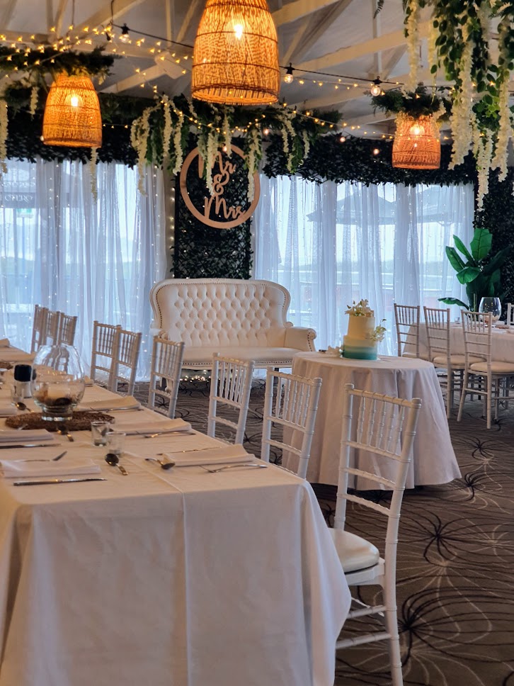 Forest Greenery door framing with open weave curtain, , Mr & Mrs 90cm timber round sign, love seat, cake table, festoon lighting, greenery ceiling bars with wisteria , White Tiffany Chairs, white linen long tables, Lotus glass centrepiece with remote candle, sand, shells, rattan placemat